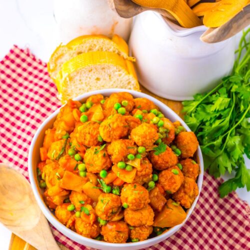 A bowl of vegetable stew with peas and meatballs sits on a red checkered cloth, surrounded by fresh parsley, bread slices, wooden spoons, and kitchen jars on a wooden board.