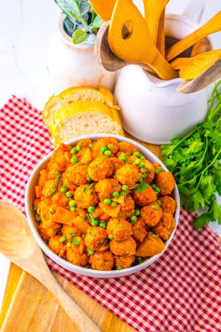 A bowl of vegetable stew with peas and meatballs sits on a red checkered cloth, surrounded by fresh parsley, bread slices, wooden spoons, and kitchen jars on a wooden board.