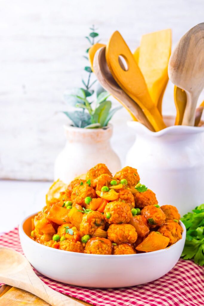 A white bowl filled with a colorful dish of saucy vegetables and golden-brown tater tots, garnished with green peas, sits on a red-checkered cloth. Wooden utensils and a plant are in the background.