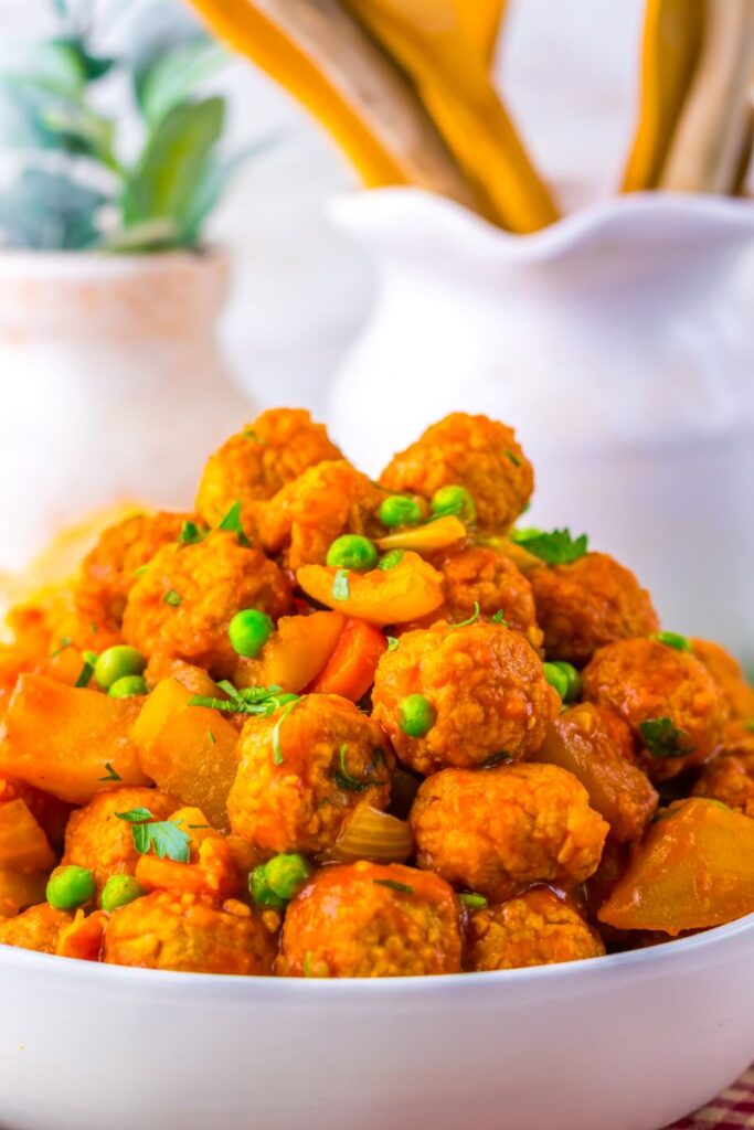 A close-up of a bowl filled with bright orange vegetable curry and meatballs, garnished with green peas and herbs. Blurred kitchen utensils and a plant are visible in the background.