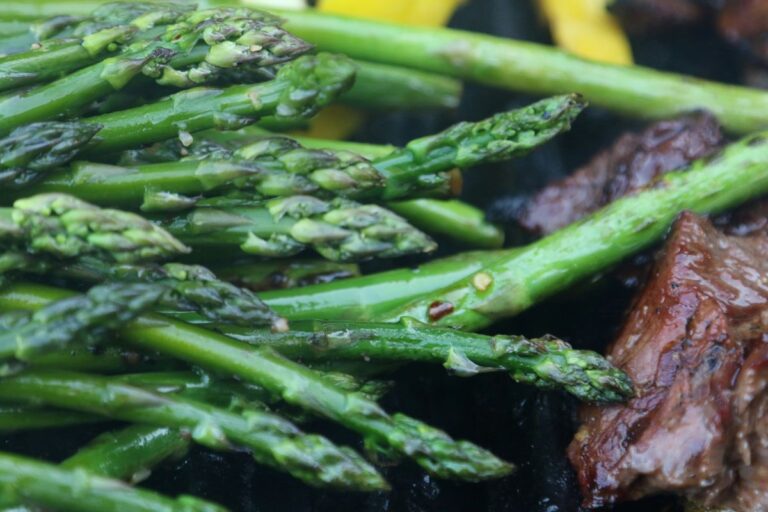 Close-up of fresh asparagus spears on a grill next to a piece of grilled meat, with some yellow vegetables visible in the background. The asparagus appears lightly seasoned.