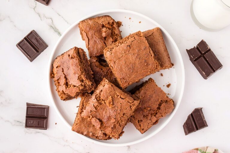 A white plate with six brownies, some with cracked tops, sits on a marble surface. Pieces of dark chocolate are scattered around the plate, and a glass of milk is partially visible in the top right corner.