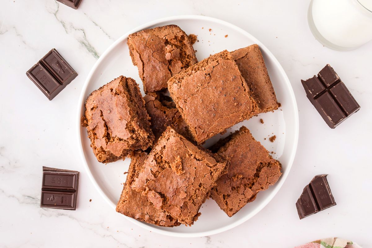 A white plate with six brownies, some with cracked tops, sits on a marble surface. Pieces of dark chocolate are scattered around the plate, and a glass of milk is partially visible in the top right corner.