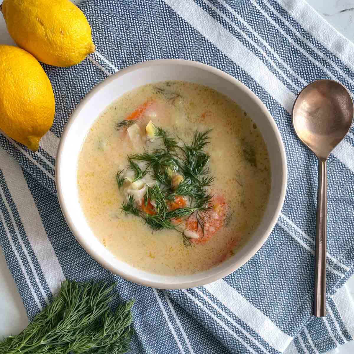 A bowl of creamy soup with herbs, chunks of vegetables, and dill garnish sits on a blue striped cloth next to a copper spoon, two lemons, and a small bunch of fresh dill.
