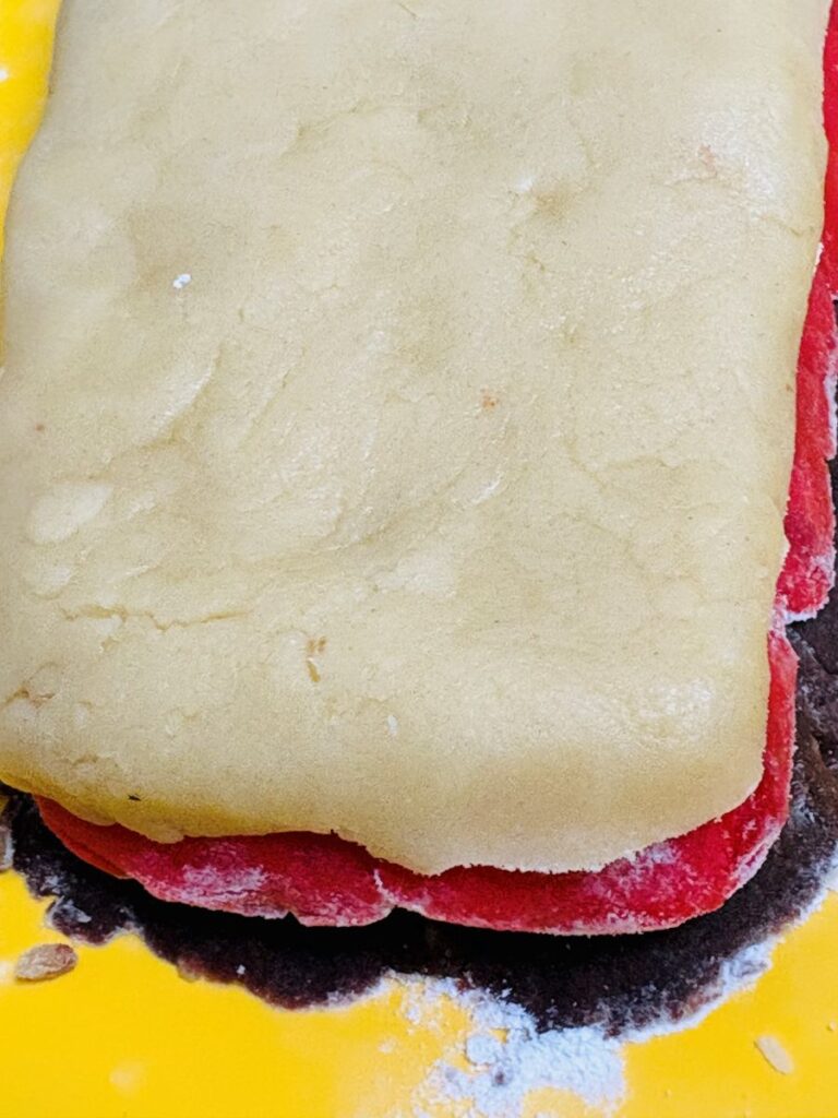 A close-up of a layered dough, with a thick layer of plain dough on top of a bright red layer, on a yellow surface lightly dusted with flour.