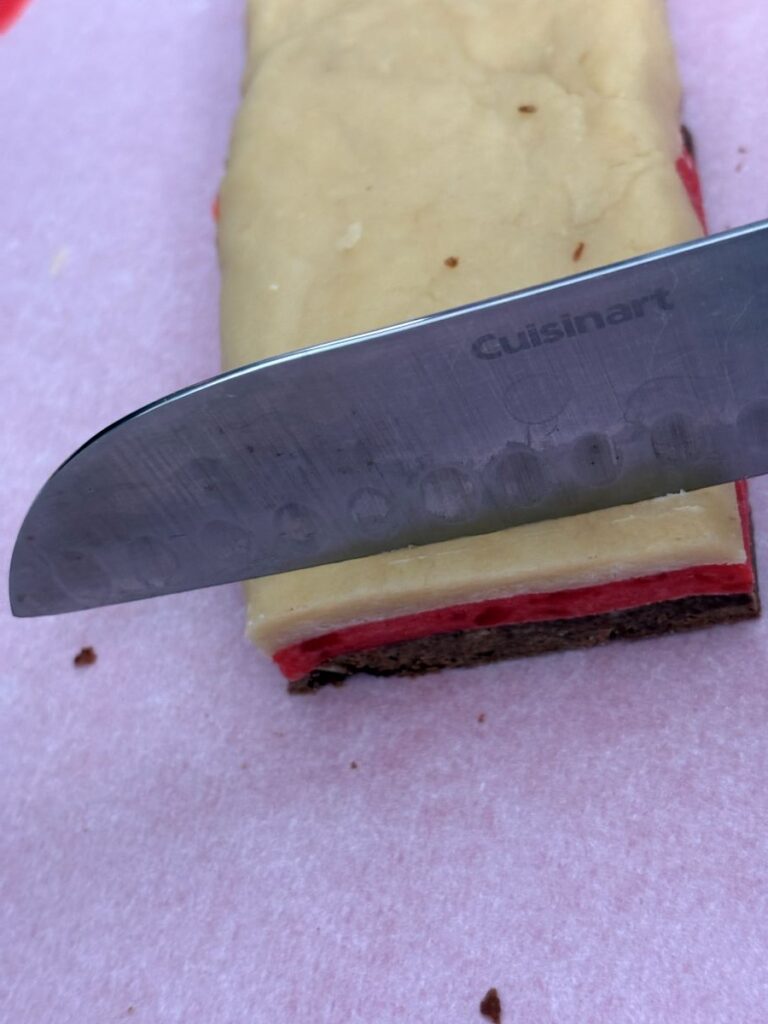 A close-up of a knife slicing through a rectangular layered cookie with brown, red, and yellow layers, resting on pink parchment paper. The knife blade is labeled “Cuisinart.”.