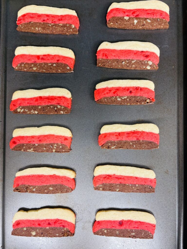Rectangular cookies with three colorful layers—white, red, and brown with nut pieces—are arranged in neat rows on a dark baking tray.