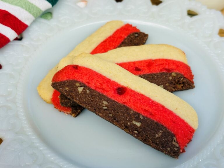 Three rectangular cookies with layers of white, red, and brown dough are arranged on a white plate. The brown layer appears to have small nut pieces. A striped cloth is partially visible in the background.