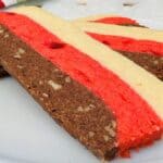 Three rectangular layered cookies on a white plate, each with chocolate, red, and vanilla-colored stripes. The chocolate layer contains small nut pieces.