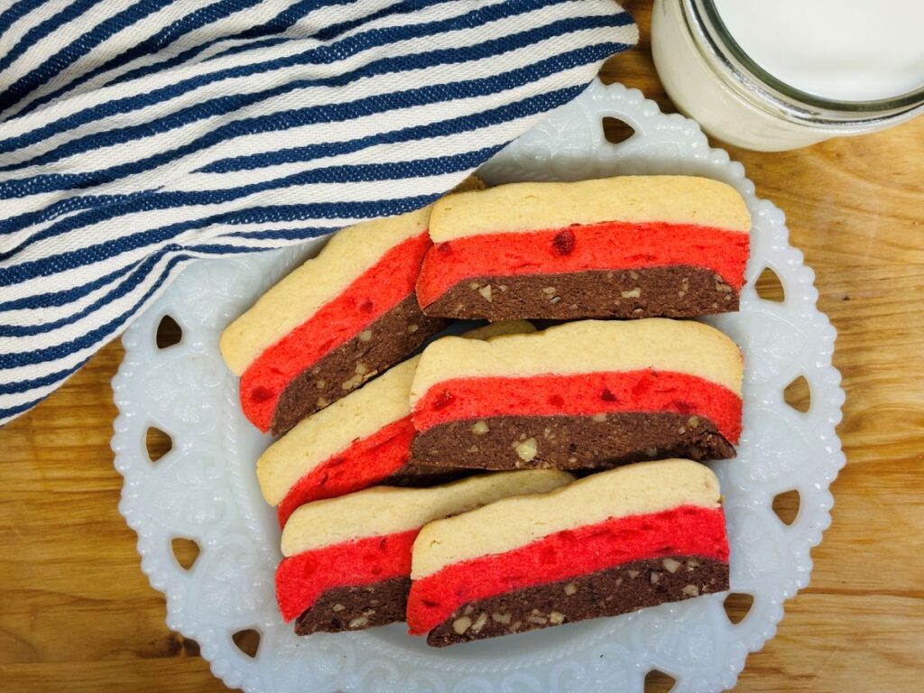 A plate of layered cookies with red, brown, and beige sections sits on a white decorative plate next to a striped cloth and a glass of milk on a wooden surface.