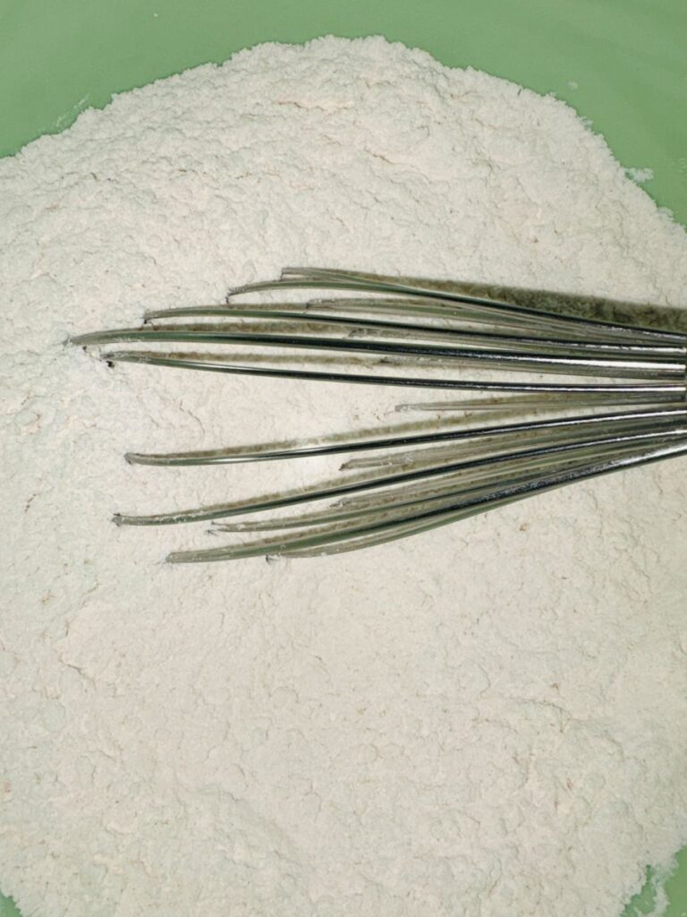 A metal whisk rests on a pile of white flour in a green mixing bowl, ready for baking preparation.