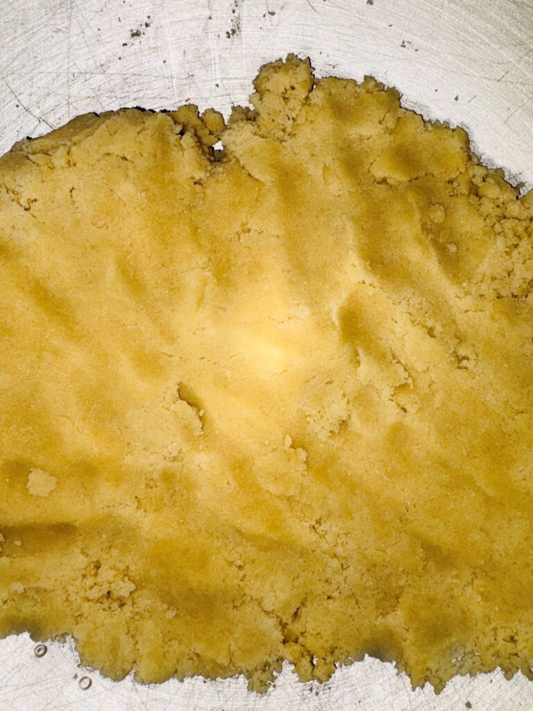 A close-up of yellow, crumbly dough spread on a metal surface, with visible texture and uneven edges.