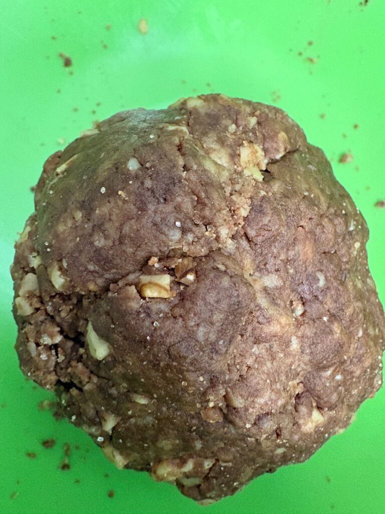 A close-up of a ball of brown cookie dough with visible chunks of nuts, placed on a bright green surface with some crumbs scattered around.