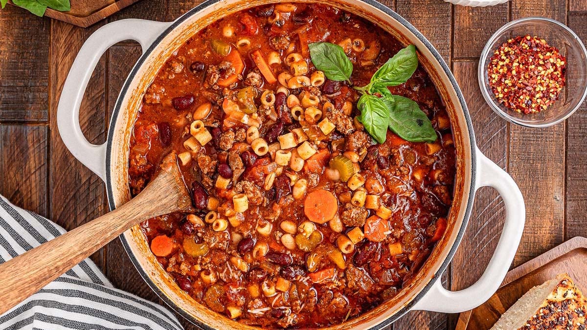 A pot of hearty minestrone soup with pasta, beans, carrots, and ground meat in a tomato broth, topped with fresh basil. A wooden spoon rests inside the pot, and bowls of red pepper flakes and bread are nearby.