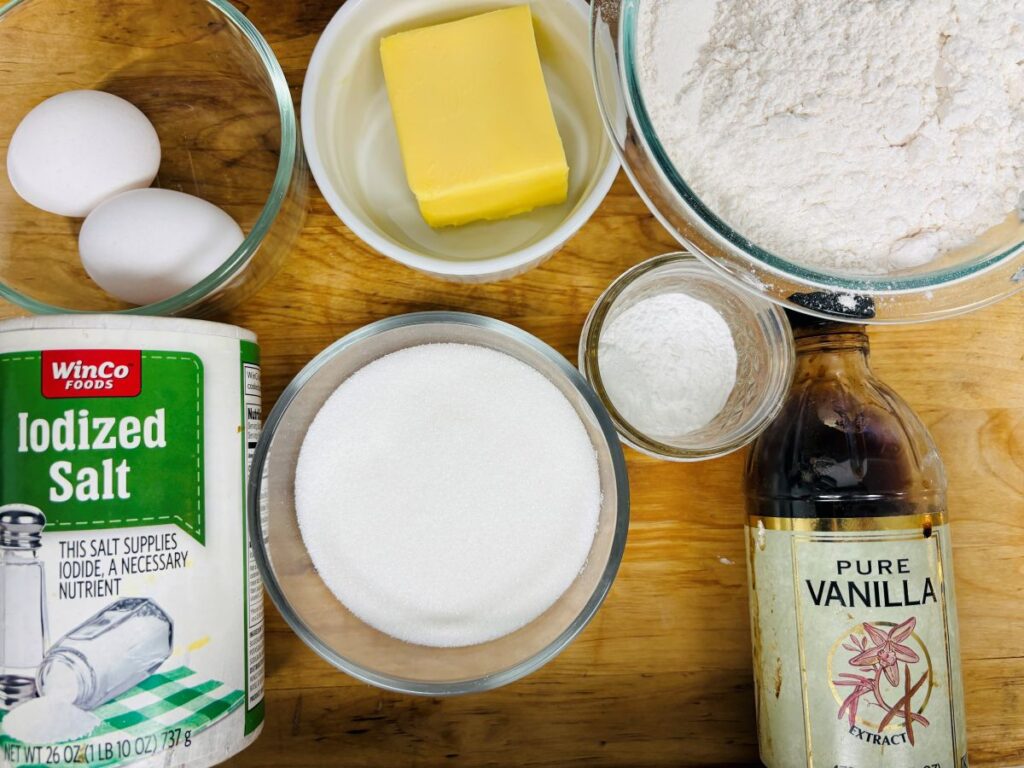 Baking ingredients on a wooden surface: two eggs in a bowl, a stick of butter, flour, baking powder, vanilla extract, granulated sugar, and a container of iodized salt.