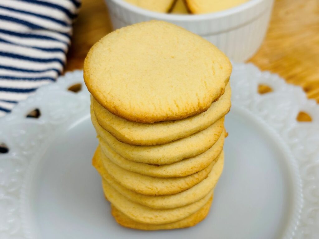 A stack of round, golden-brown sugar cookies sits on a white plate with a decorative edge. A bowl with more cookies and a striped cloth are in the background.