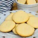 A white plate with several golden brown shortbread cookies sits in front of a striped cloth; more cookies are in a white ramekin in the background.