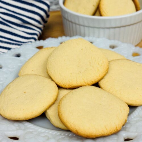 A white plate with several golden brown shortbread cookies sits in front of a striped cloth; more cookies are in a white ramekin in the background.