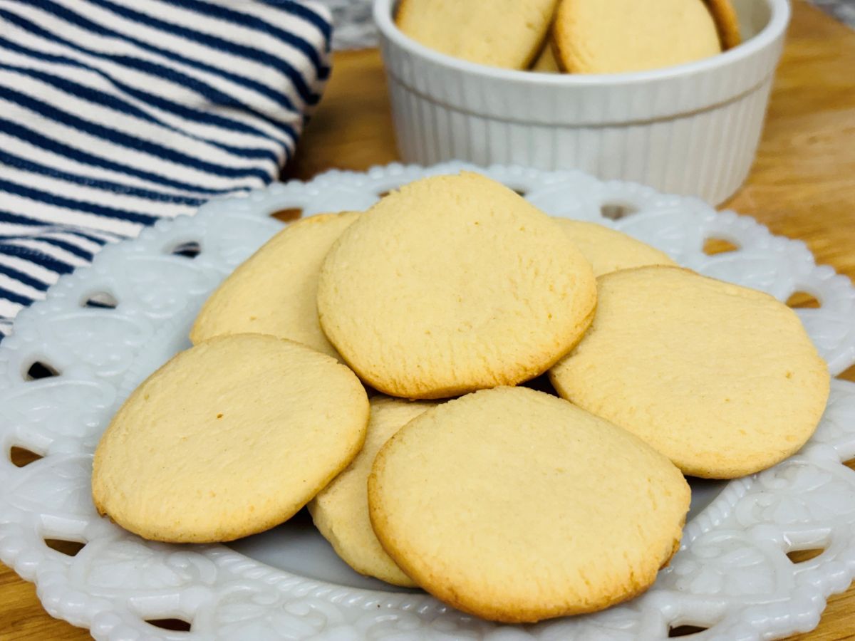 A white plate with several golden brown shortbread cookies sits in front of a striped cloth; more cookies are in a white ramekin in the background.