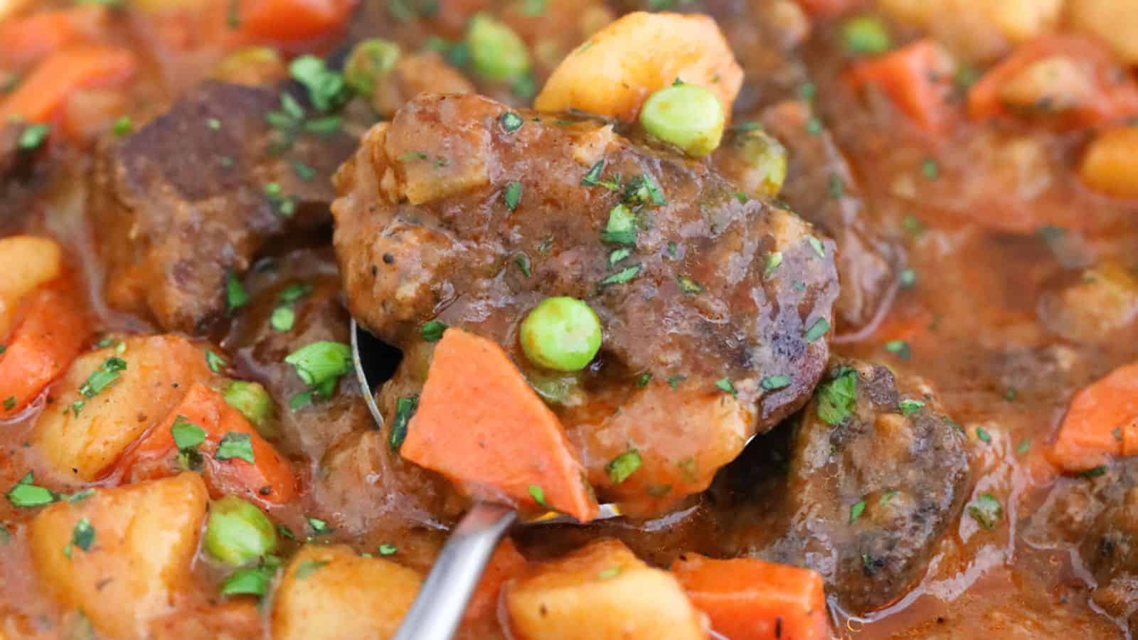 A close-up of a hearty beef stew with chunks of beef, potatoes, carrots, green peas, and garnished with chopped parsley in a rich, brown gravy. A spoon is lifting a serving from the stew.