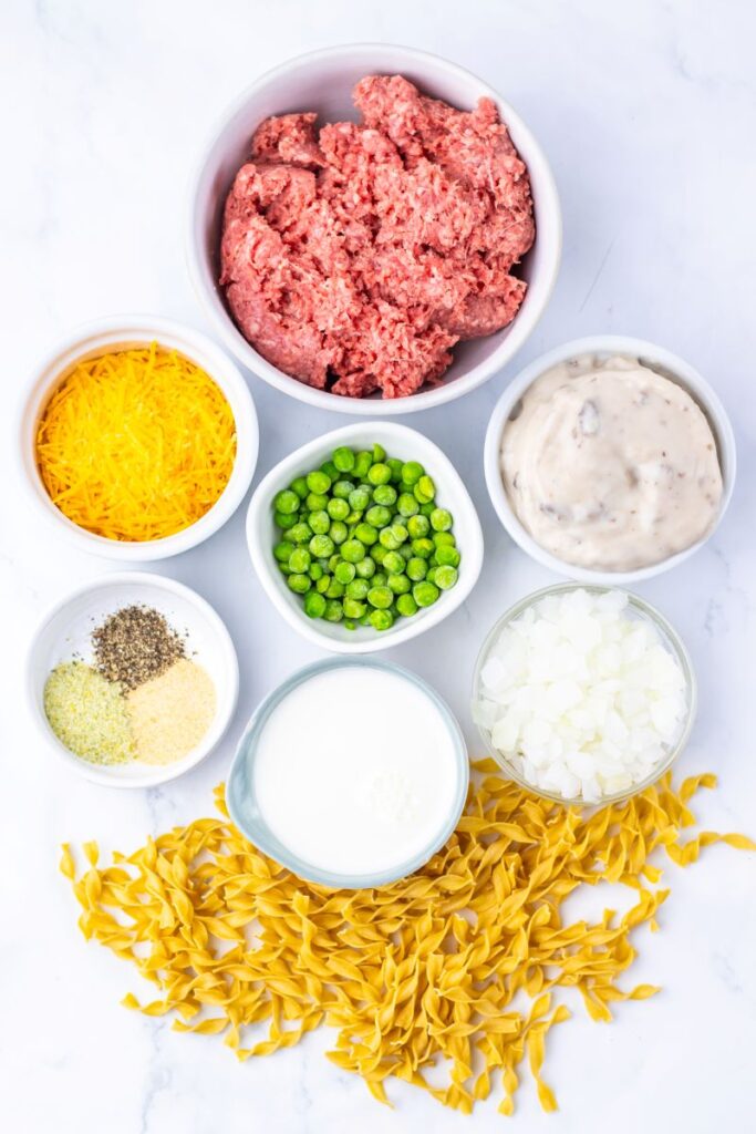 A flat lay of ingredients in bowls and on a white surface: ground beef, shredded cheddar cheese, peas, cream of mushroom soup, chopped onions, seasonings, milk, and uncooked egg noodles.