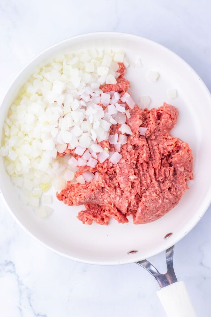 A white skillet containing raw ground meat and chopped onions, both uncooked and placed side by side on a white surface.