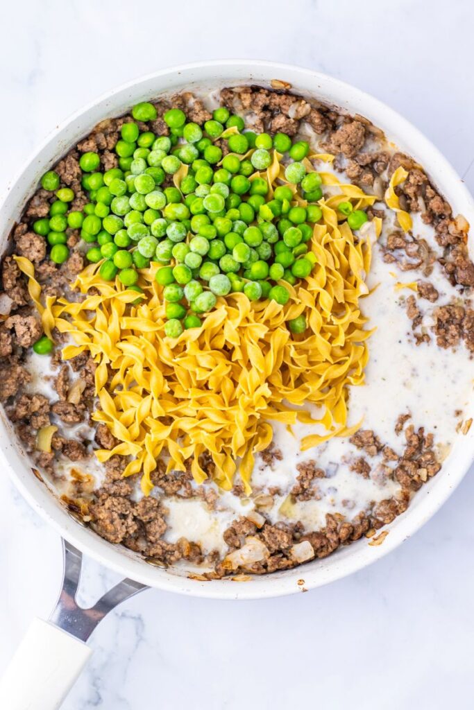 A white skillet contains cooked ground beef, egg noodles, green peas, and a creamy sauce, all partially mixed together on a marble surface.