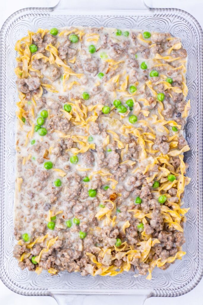 A casserole dish filled with a mixture of cooked ground beef, egg noodles, green peas, and a creamy sauce, spread evenly and ready to serve.