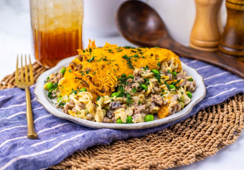 A plate of cheesy casserole with ground meat, noodles, peas, and a golden melted cheese topping sits on a woven mat, next to a fork and a glass of iced drink. Wooden utensils and salt and pepper shakers are in the background.