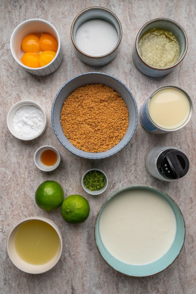 A flat lay of Key lime pie ingredients: egg yolks, sugar, butter, graham cracker crumbs, sweetened condensed milk, baking powder, vanilla extract, lime zest, limes, lime juice, and heavy cream on a light surface.