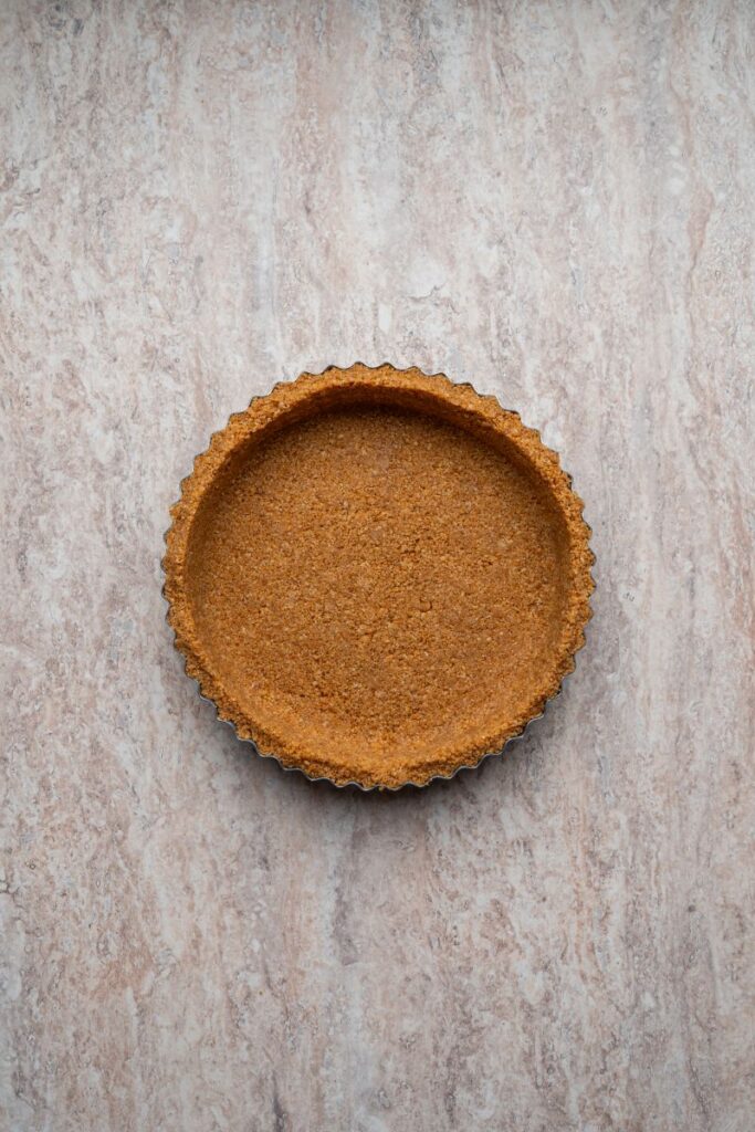 A round tart pan filled with a pressed graham cracker crust sits on a light, marbled countertop. The crust has an even, golden-brown color and slightly scalloped edges.