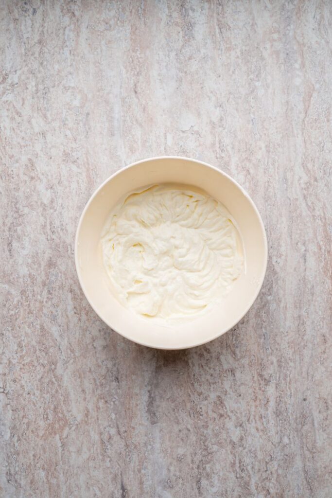 A white mixing bowl filled with creamy, whipped mixture sits on a light marbled countertop, viewed from above.