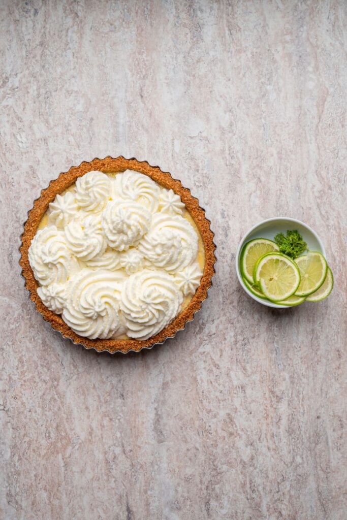 A key lime pie topped with swirls of whipped cream sits on a light stone surface next to a small bowl containing lime slices and a sprig of parsley.