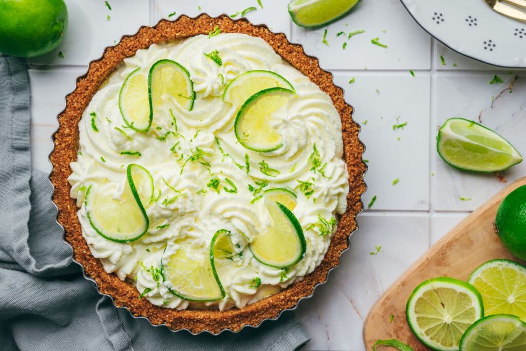A key lime pie topped with whipped cream, lime slices, and lime zest sits on a white tiled surface with fresh limes and lime wedges nearby. A gray napkin and a cutting board with lime slices are also visible.