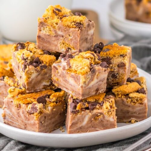 A plate stacked with fudge squares topped with crumbled cookies and chocolate chips, set on a gray cloth with a blurred background including a glass of milk and a bowl of white candies.
