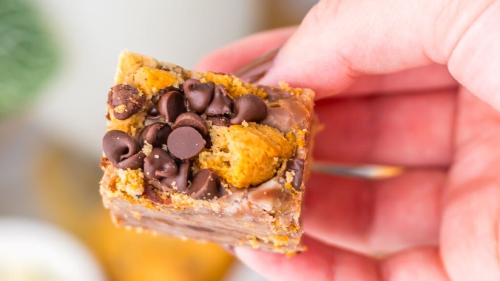 A close-up of a hand holding a dessert bar topped with chocolate chips and cookie pieces. The treat has a layered, crumbly texture and appears rich and sweet.