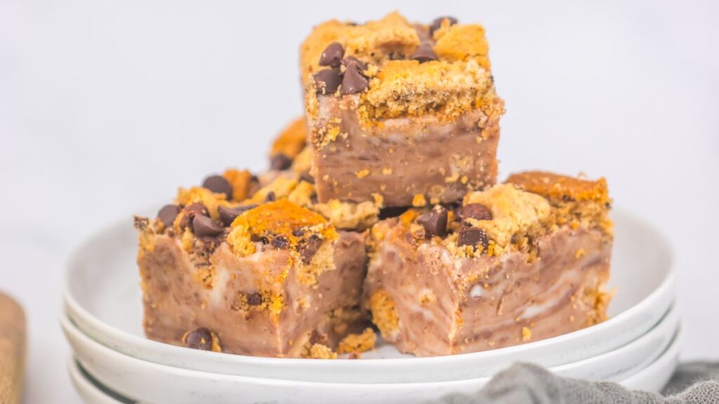 Three pieces of chocolate chip cookie dough fudge are stacked on a white plate. The fudge is creamy with chocolate chips and cookie pieces visible. The background is light and out of focus.