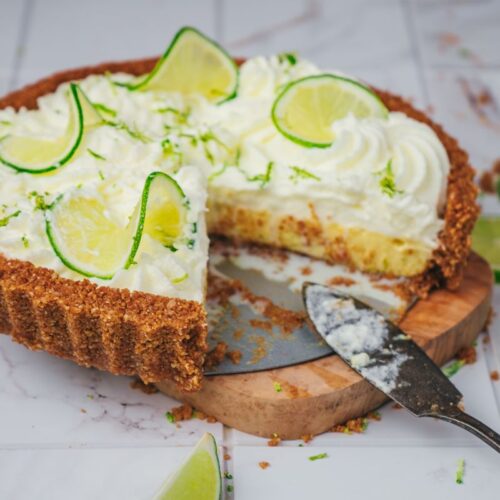 A key lime pie topped with whipped cream and lime slices sits on a wooden board. A slice has been cut and a pie server rests nearby, with crumbs and lime wedges scattered around.