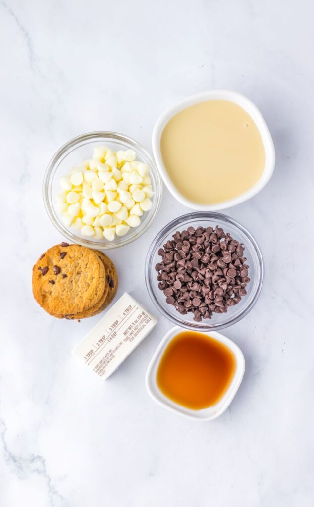 Five ingredients on a white surface: chocolate chip cookies, a stick of butter, a bowl of white chocolate chips, a bowl of mini chocolate chips, a bowl of sweetened condensed milk, and a small bowl of vanilla extract.