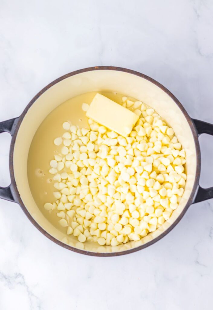 A black pot containing sweetened condensed milk, white chocolate chips, and a stick of butter on a white marble surface.