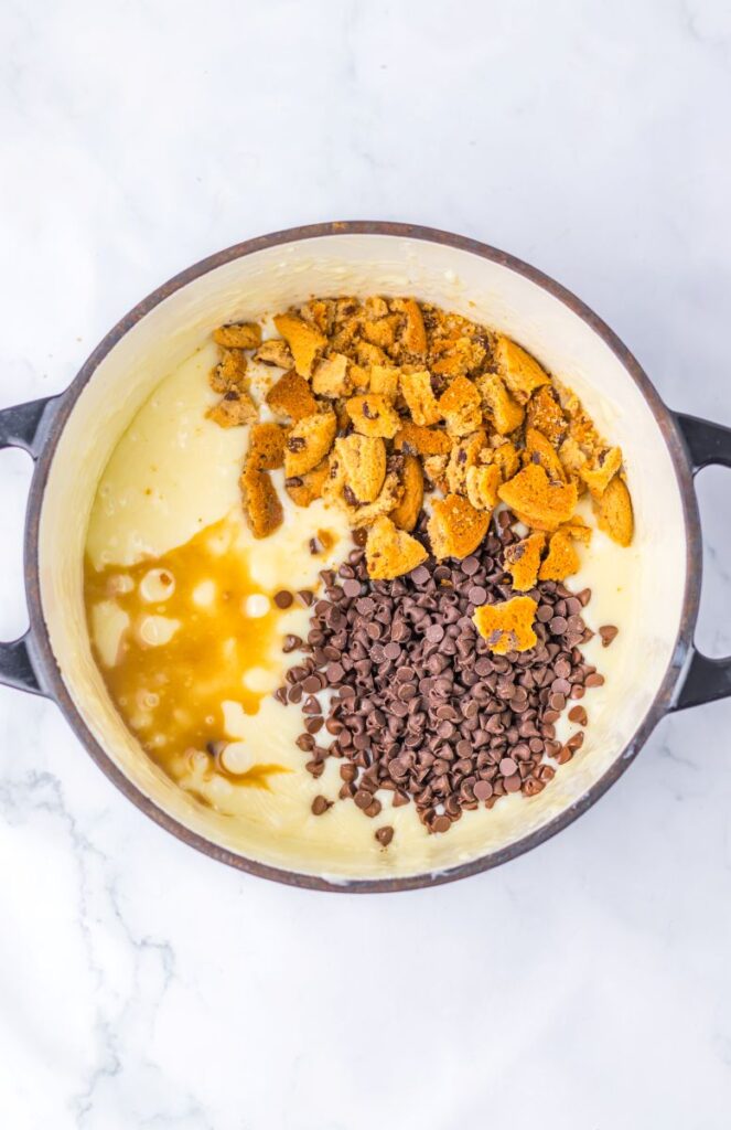 A large mixing bowl filled with creamy batter, topped with broken cookie pieces and chocolate chips, sits on a white marble surface.