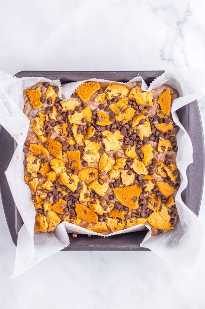 A square baking pan lined with parchment paper holds a dessert topped with broken chocolate chip cookies and chocolate chips on a light marble surface.