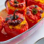 A glass baking dish filled with roasted red bell peppers stuffed with a yellow rice mixture, some peppers slightly charred on top. The dish is placed on a white surface next to a gray-edged plate.