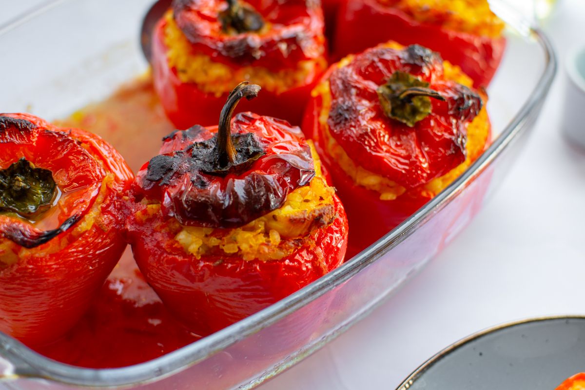 A glass baking dish filled with roasted red bell peppers stuffed with a yellow rice mixture, some peppers slightly charred on top. The dish is placed on a white surface next to a gray-edged plate.