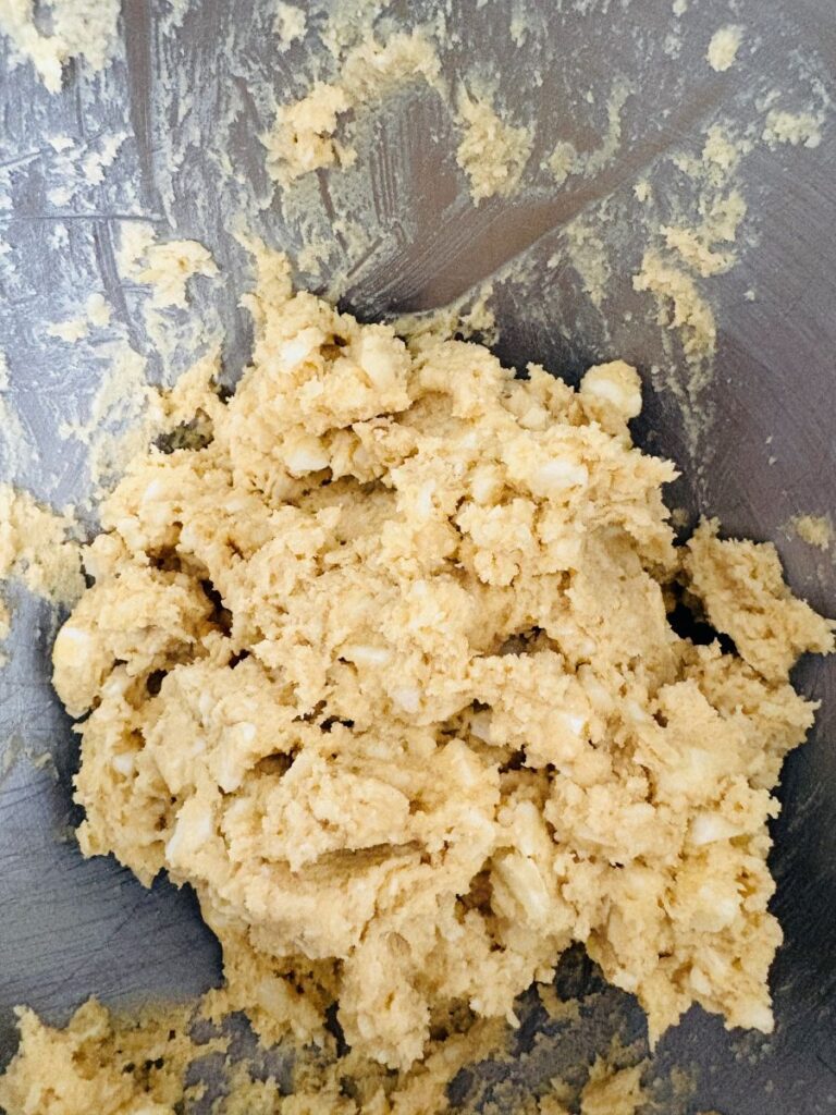 A close-up view of raw cookie dough with visible chunks, spread in a metal mixing bowl. The dough appears thick and slightly crumbly.