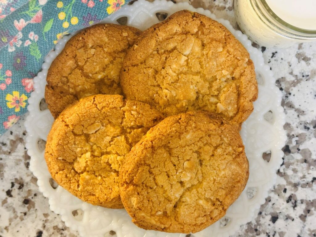 Four golden-brown cookies are arranged on a white decorative plate, placed on a speckled countertop. A glass of milk and a colorful floral napkin are visible near the plate.
