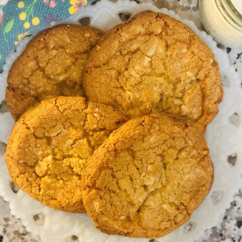 Four golden-brown cookies are arranged on a white decorative plate, placed on a speckled countertop. A glass of milk and a colorful floral napkin are visible near the plate.