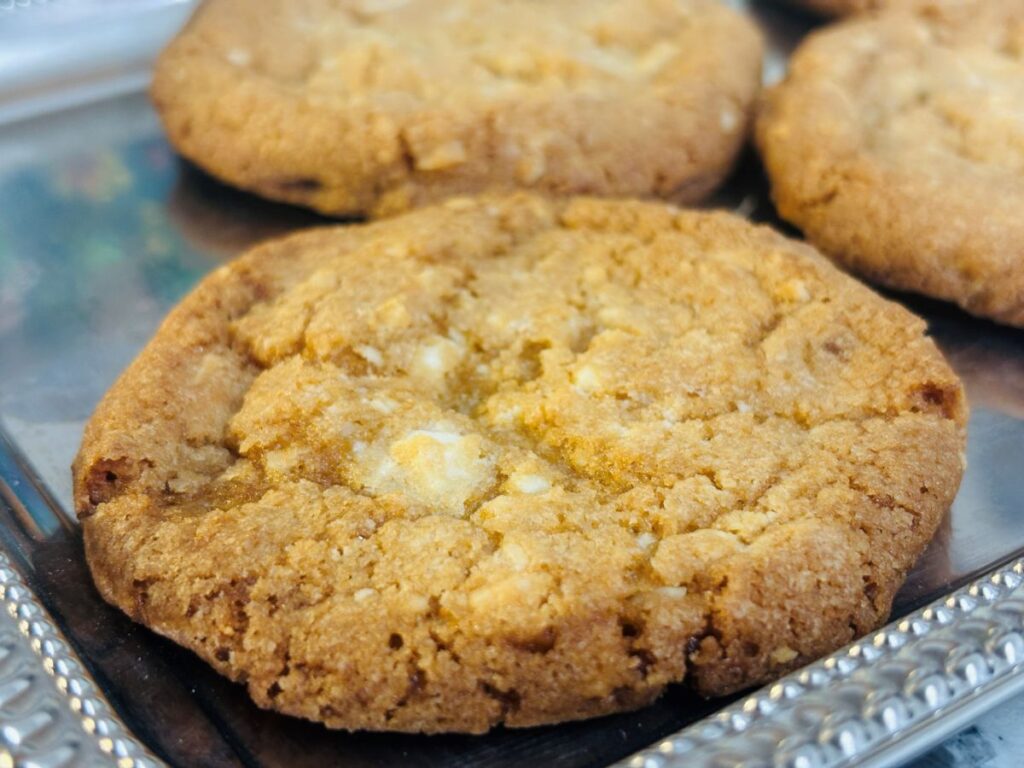 Close-up of a golden brown cookie with visible white chocolate chunks, placed on a silver tray with other similar cookies in the background. The cookie has a slightly crisp and crumbly texture.