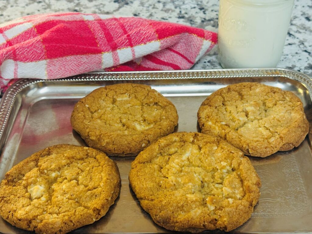 Four large golden brown cookies on a silver tray, with a red and white checkered cloth and a glass of milk in a mason jar in the background on a speckled countertop.