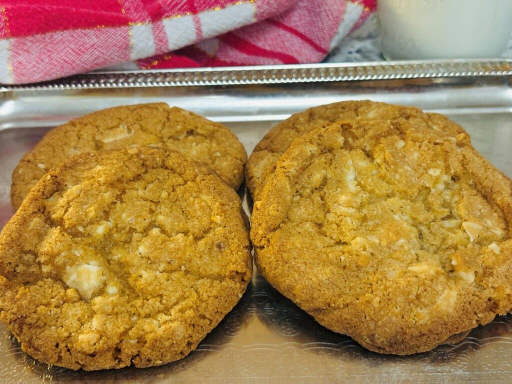 Four golden brown cookies with white chocolate chunks are arranged on a silver tray. A red and white checkered towel and a glass of milk are visible in the background.